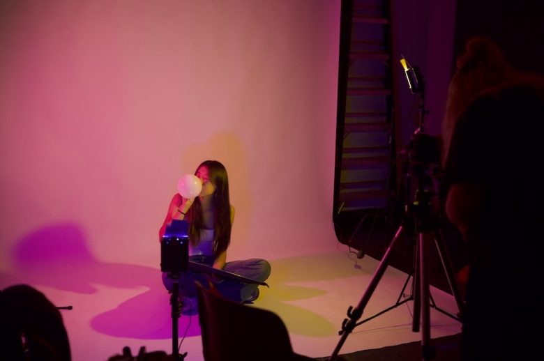 A Tisch Summer High School Photography student poses in front of a camera and white portrait background that is lit by a rose pink light. The student is blowing up a white latex balloon. Two cameras on tripods are also shown in the image.